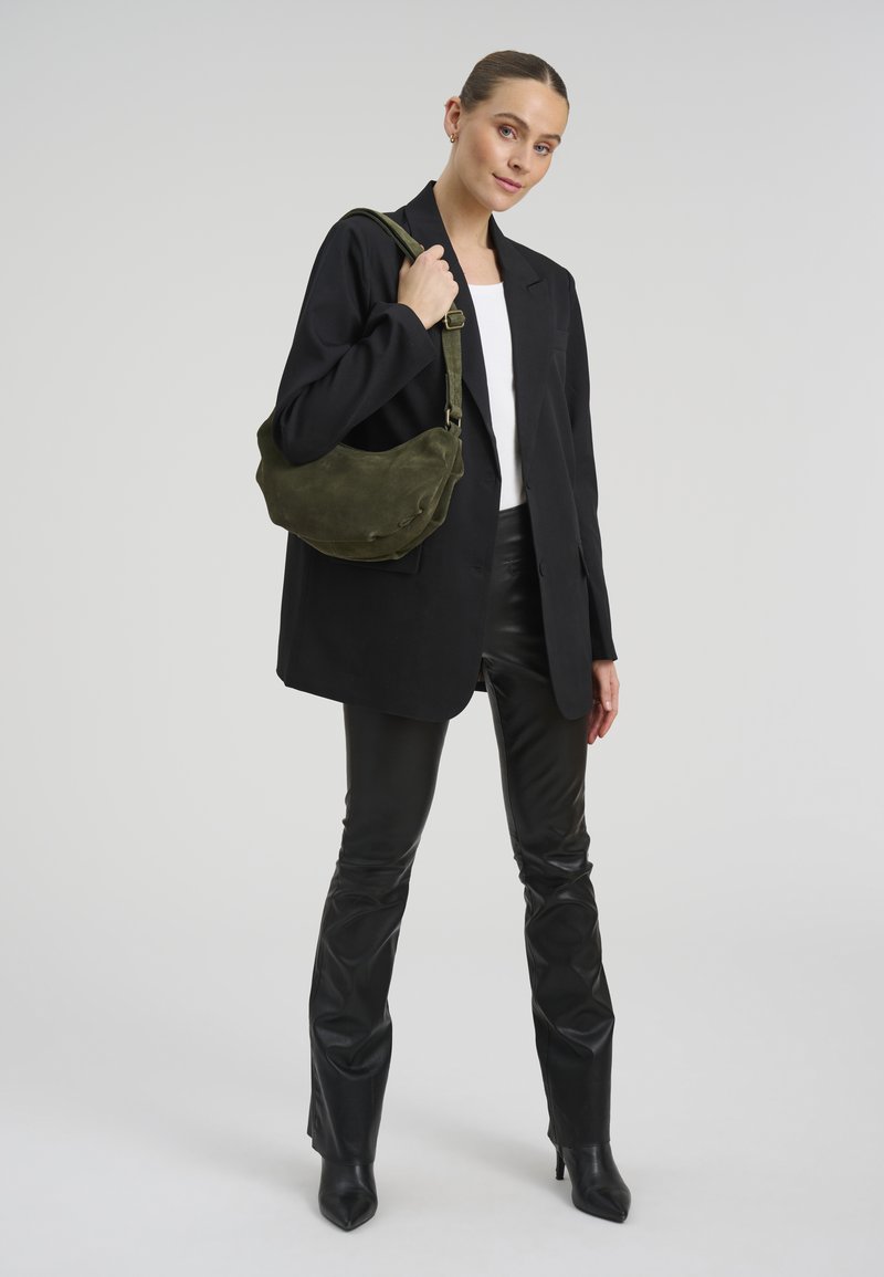 Femme portant un blazer noir, un haut blanc, un pantalon en cuir noir, des bottines à talons noires, tenant un sac épaule en daim vert olive sur fond gris.