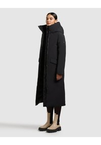 Lang sort dunjakke med høj krave, lynlås foran og store sidelommer. Modellen har beige ankelstøvler på med en tyk sål.