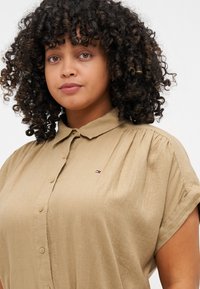 Khaki kurzärmliges Hemd mit Knöpfen, leichtes Material, mit kleinem Logo auf der linken Seite. Glatte Textur, lässige Passform.