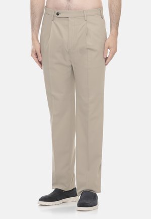 Pantaloni beige plissettati indossati da una persona in piedi con il petto nudo, abbinati a scarpe slip-on nere con suole bianche.