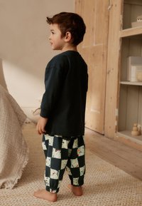 Maglia a maniche lunghe verde scuro abbinata a pantaloni a fantasia con quadrati bianchi e neri e spruzzi colorati. Piedi scalzi, pavimento in legno chiaro.