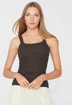 Giovane donna con lunghi capelli biondi che indossa una canotta attillata marrone scuro e una gonna chiara, in piedi con le mani intrecciate.