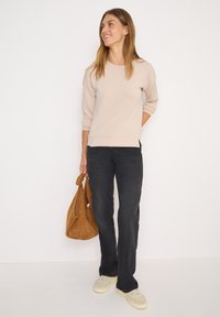 Sweatshirt beige clair à manches trois-quarts, associé à un jean foncé et des baskets beiges, tenant un sac fourre-tout en daim marron.