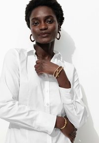 Chemise blanche à boutons, bracelets dorés en couches et grandes boucles d'oreilles dorées. Un tissu lisse et des accents métalliques brillants mettent en valeur le look.