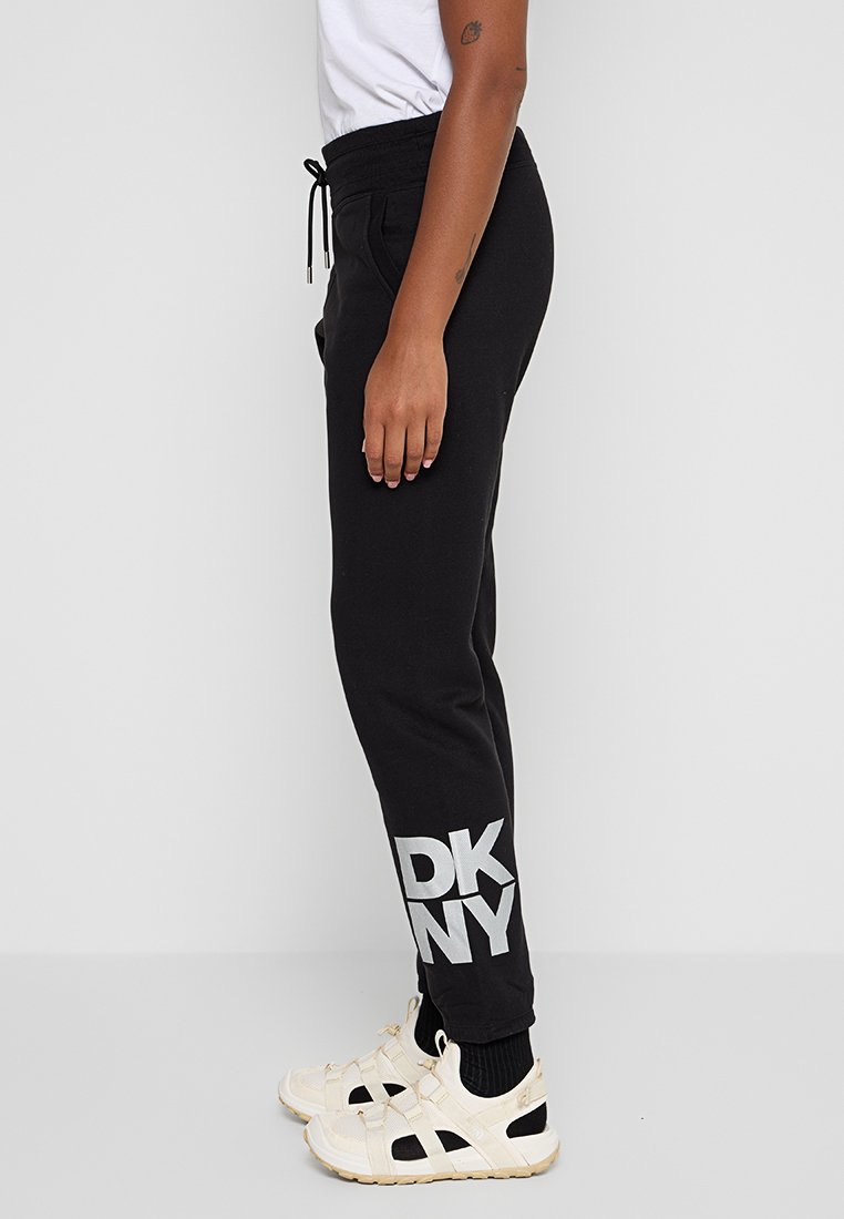 Persona che indossa pantaloni sportivi neri DKNY, scarpe da tennis bianche, calze nere e una maglietta bianca su uno sfondo chiaro semplice.