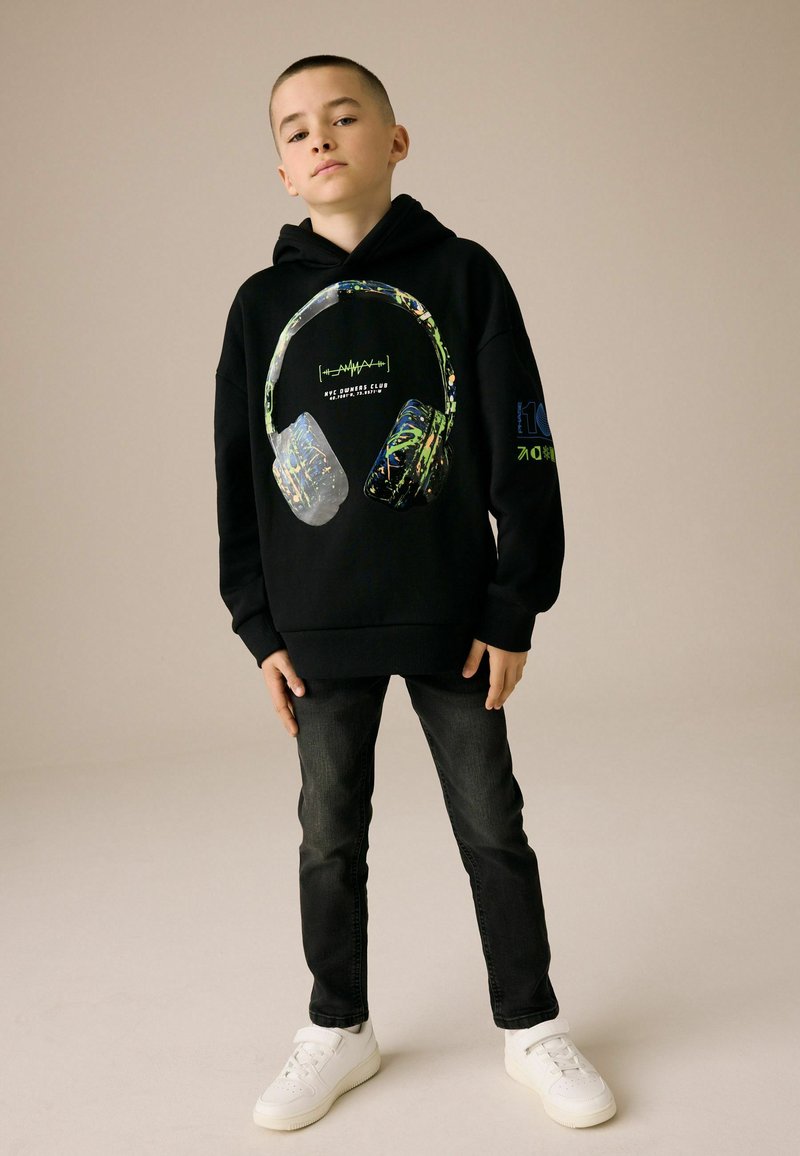 Zwarte hoodie met een grafisch ontwerp van koptelefoons in levendige kleuren. Gecombineerd met donkere jeans en witte sneakers, wat een casual look creëert.
