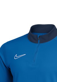 Maglia atletica blu a mezza zip realizzata in tessuto leggero. Presenta un colletto alto, dettagli in contrasto blu navy e un logo bianco sul lato sinistro del petto.