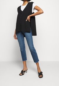 Top de punto negro sin mangas con un escote en V profundo, superpuesto a una camisa blanca, combinado con jeans azul cortos y sandalias de tacón negras.