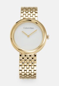 Calvin Klein zlatni sat s okruglim broj�čanikom i bijelim pozadinom. Ima metalnu narukvicu i dva zlatna kazaljka za sate.