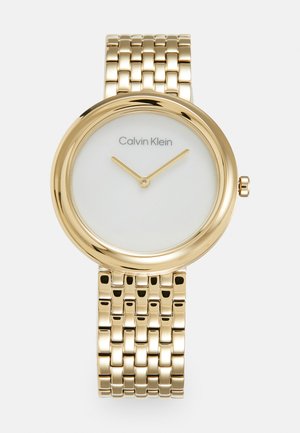 Montre Calvin Klein dorée avec un cadran rond et un fond blanc. Dispose d'un bracelet en maillons métalliques et de deux aiguilles dorées pour les heures.