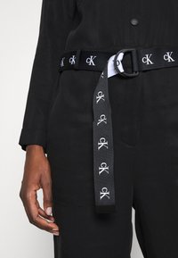 Ceinture en tissu noir avec une texture lisse, dotée d'une boucle noire et d'un logo "ck" brodé en blanc. La ceinture a un design de sangle plate.