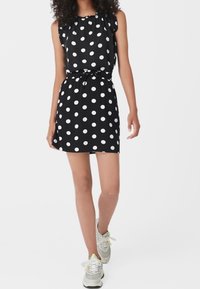 Abito nero senza maniche con pois bianchi, scollatura arricciata, vita cintrata e gonna svasata. Indossato con sneakers grigie.