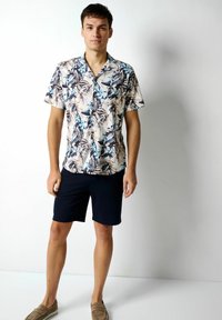 Kurzärmliges, kragenbekleidetet Hemd mit floralem Muster in Beige-, Blau- und Brauntönen, kombiniert mit dunkelblauen Shorts und hellbraunen Schuhen.