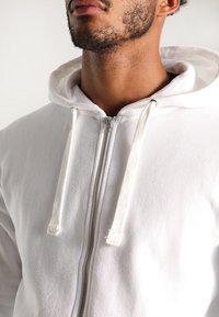 Sudadera blanca con cremallera y textura en el tejido, con capucha ajustable y detalles de cremallera plateados, que muestra un diseño limpio y minimalista.
