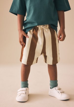 Bambino in piedi con pantaloncini a righe marroni e beige, calzini verdi, scarpe da ginnastica bianche con velcro e una maglietta oversize color verde acqua.