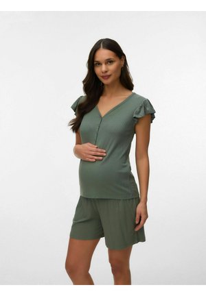 Zwangere vrouw met lang donker haar, gekleed in een olijfgroene korte mouw top en bijpassende korte broek, staand tegen een effen lichte achtergrond.