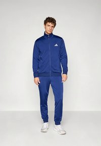 Tumši zils sporta apģērbu komplekts no gludas, vieglas auduma. Komplektā ietilpst jostas rāvējslēdzēja jaka un šauras bikses, abām ar baltām logotipa detaļām.