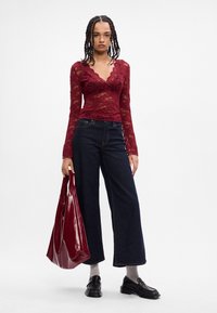 Top de renda bordô de mangas longas com decote em forma de onda, combinado com calças largas de ganga escuras e uma mala de mão brilhante burgundy. Sapatos pretos e meias cinzas.