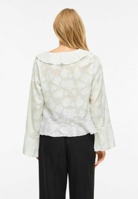 �Ženska z dolgimi blond lasmi, ki nosi belo prosojno cvetlično bluzo z nabranim ovratnikom in črne hlače, prikazana od zadaj.