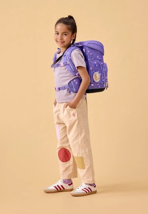 Purple backpack with polka dots and a character patch, worn with beige pants featuring colorful fabric patches and white sneakers with red stripes.
