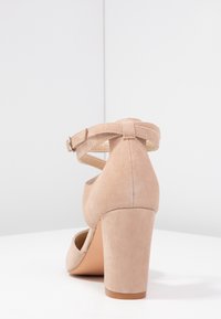 Beige mockask högklackad sko med rund tå och ankelrem, med klack i blockform och en texturerad yta.