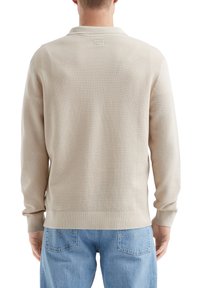 Beige Strickpullover mit einem strukturierten Muster, langen Ärmeln und einem Kragen. Endet auf Taillenhöhe und wird mit hellblauen Jeans kombiniert.