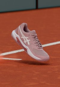 Jasnoróżowy but sportowy z siateczkową cholewką, białymi akcentami i teksturowaną podeszwą, umieszczony nad kortem ziemnym, z unoszącymi się wokół drobinami kurzu.