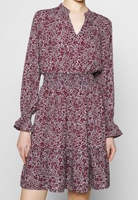 Blommig klänning med ett vinrött och vitt mönster, v-ringning, lång ärm med volanger och smockad midja med en lager-på-lager kjol.