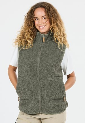 Vrouw met krullend haar die lacht, gekleed in een grijze fleecebodywarmer met rits over een wit T-shirt met korte mouwen en beige broek, handen in de vestzakken.