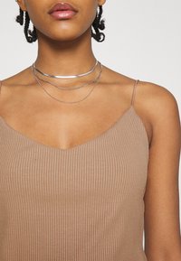 Débardeur marron à côtes avec un col en V, mettant en valeur trois colliers superposés en argent de différentes épaisseurs et textures, sur un fond neutre.