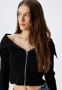 Czarny prążkowany krótki sweter z szerokim dekoltem typu off-shoulder oraz srebrnym zapięciem na zamek. Charakteryzuje się dopasowanym krojem i długimi rękawami.
