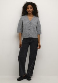 Pilkas megztas kardiganas su trimis gėlių formos sagomis, trumpomis rankovėmis, V formos iškirpte, derinamas su juodais aukštos liemens džinsais ir juodais batais.