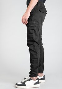 Pantalones cargo negros de denim, con un ajuste slim, múltiples bolsillos y detalles de costura visibles. Combinados con zapatillas blancas y negras.
