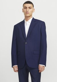 Blazer ajustado color azul marino con solapas de muesca, cierre de un solo botón, dos bolsillos frontales y textura de tela suave. Camisa blanca debajo.