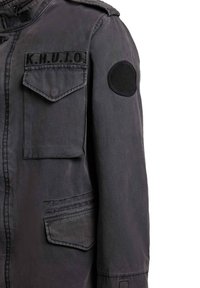 Veste gris foncé avec des poches à rabat, un écusson sur la poitrine gauche portant l'inscription "K.H.U.J.O," et un écusson rond noir sur le haut de la manche gauche.
