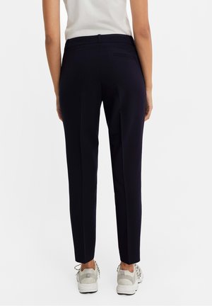 Pantalon bleu marine ajusté, à coupe élégante, présentant une texture lisse et des poches arrière discrètes, associé à des baskets de couleur claire.
