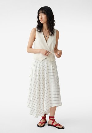 Donna con capelli neri mossi che indossa un gilet avvolgente beige senza maniche, gonna midi a righe bianche, collane a strati, anelli e sandali piatti rossi con cinturini.