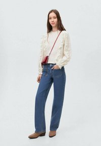 Blusa bianca a maniche lunghe con dettagli arricciati, abbinata a jeans blu a vita alta e scarpe con tacco marroni, completata da una borsa a tracolla rossa.