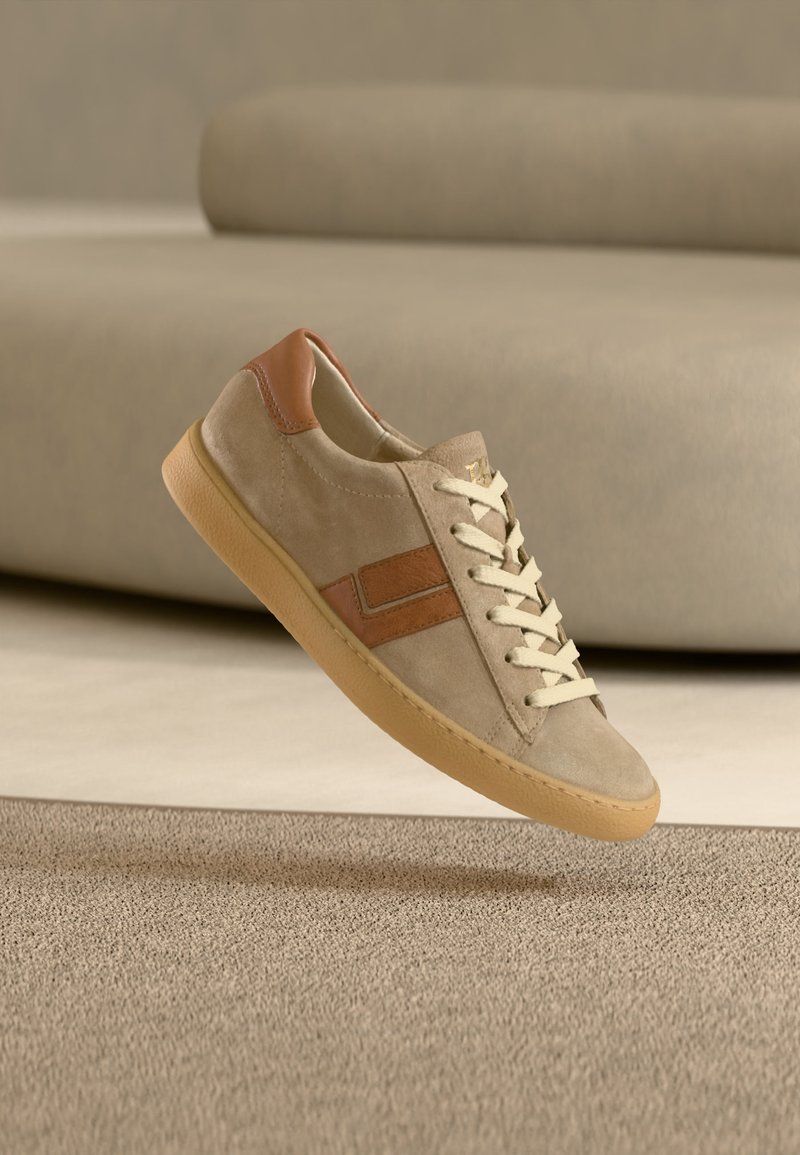 Basket en daim beige et marron avec des lacets crème et une semelle en gomme flottant au-dessus d'un tapis beige avec un canapé assorti en arrière-plan.