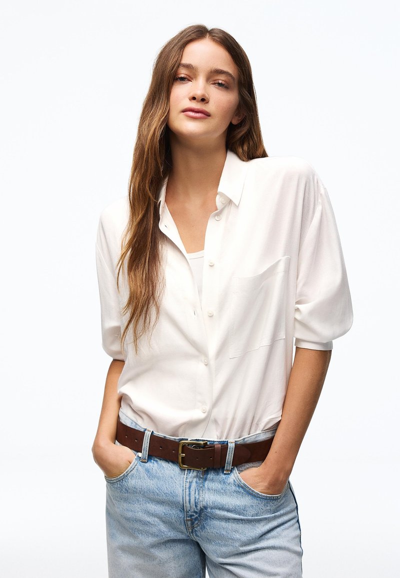 Nuori nainen, jolla on pitkät ruskeat hiukset, valkoinen napillinen paita ja vaaleansiniset farkut ruskealla vyöllä, kädet taskuissa.