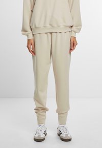 Beige joggebukser laget av mykt stoff, med en avslappet passform, elastisk midje og mansjetter. Brukt med hvite og brune joggesko.