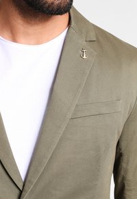 Marynarka oliwkowa o fakturowanym wykończeniu, z pojedynczą boczną kieszenią i ozdobnym przypinkiem w kształcie kotwicy na klapie, zestawiona z białym t-shirtem.