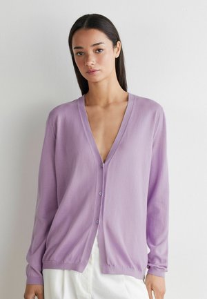 Ung kvinde med glat mørkt hår iført en lys lilla V-hals knapset cardigan og hvide bukser mod en ensfarvet hvid baggrund.