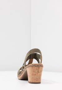 Oliivinvihreä nubukista valmistettu korokepohjainen sandaali, jossa on punottu somistus ja korkkipohja, nähtynä takaa valkoisella pinnalla ja seinätaustalla.