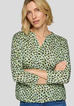 Frau mit kurzen blonden Haaren, die ein langärmliges Oberteil mit grün-blauem Leopardenmuster trägt und die Arme vor dem Körper verschränkt, vor einem schlichten Hintergrund.