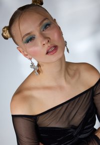 Top nero scollato realizzato in materiale trasparente, con un design arricciato. La modella indossa orecchini a forma di croce scintillanti e un trucco per gli occhi in un audace tono turchese.