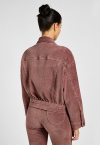 Giacca in corduroy rosa con vita aderente, maniche lunghe e collo a camicia; presenta righe verticali e dettagli cuciti.