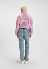 Fleecejacka i gradientlila med hög krage, märkeslogotyp på baksidan. Matchad med ljusblå jeans och vita sneakers.