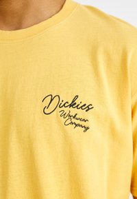 Geltona medvilninė marškinėliai su juodu siuvinėtu "Dickies Workwear Company" logotipu kairėje pusėje krūtinės, su klasikine apvalia kaklo iškirpte.