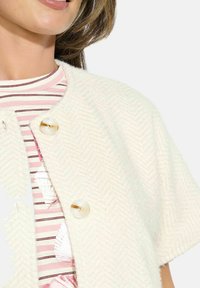 White herringbone textured cardigan with short sleeves, featuring two clear buttons. Worn over a pink striped shirt with shell prints.
